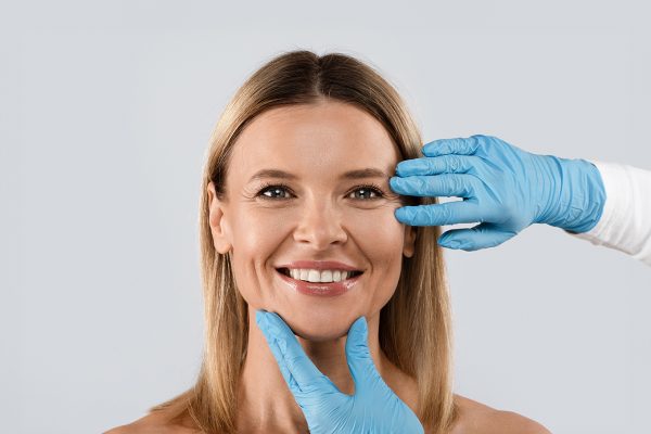 Portrait of smiling middle aged beautiful blonde woman getting prepared for plastic surgery procedures by doctor touching her face skin, isolated on grey background, copy space